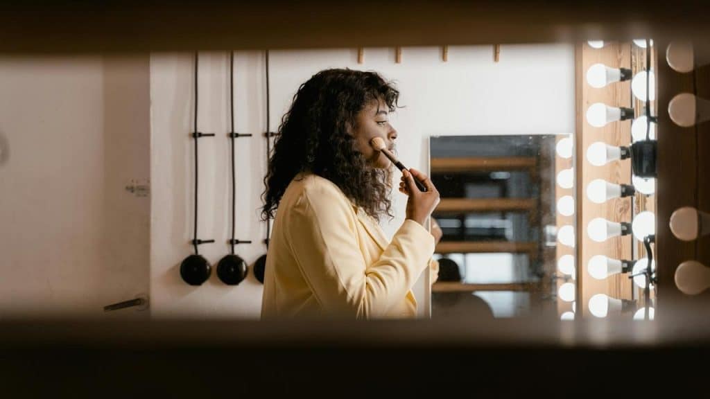 A woman applies makeup in front of a mirror surrounded by bright lights.