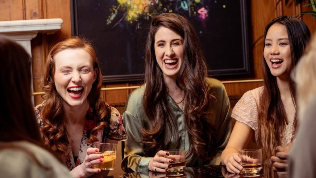 Three women sit around a dark table laughing together while holding glasses containing amber liquid.