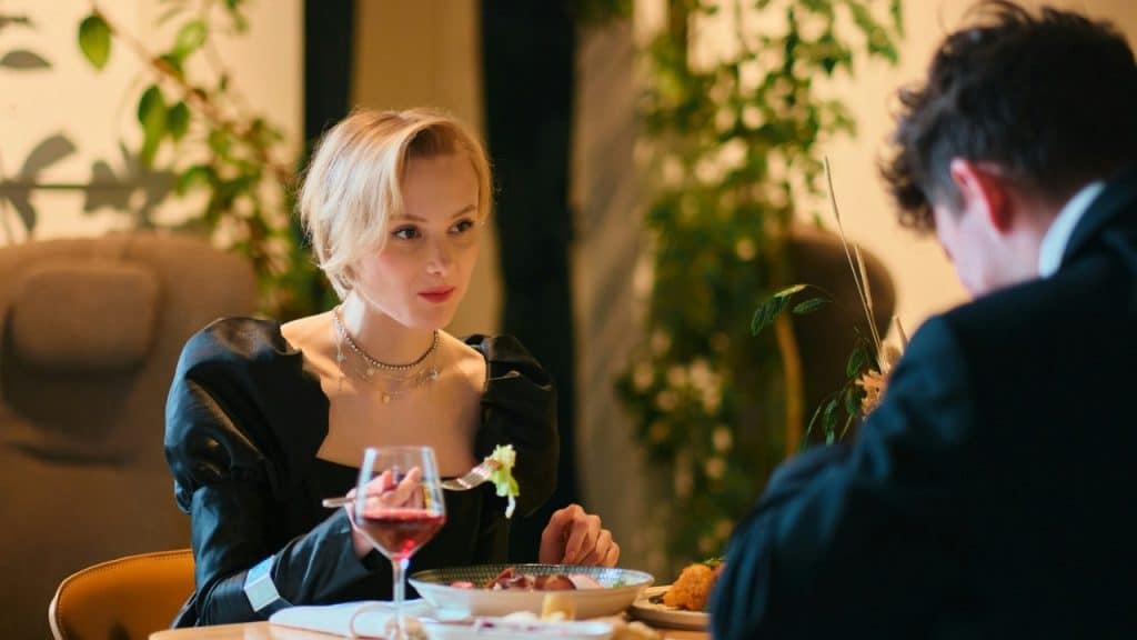 A blonde woman in a black dress sits at a dinner table looking at a man.