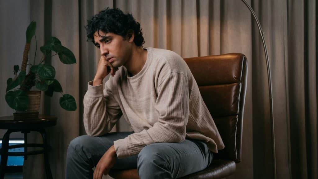 A man with dark curly hair sits in a leather chair looking down and away.