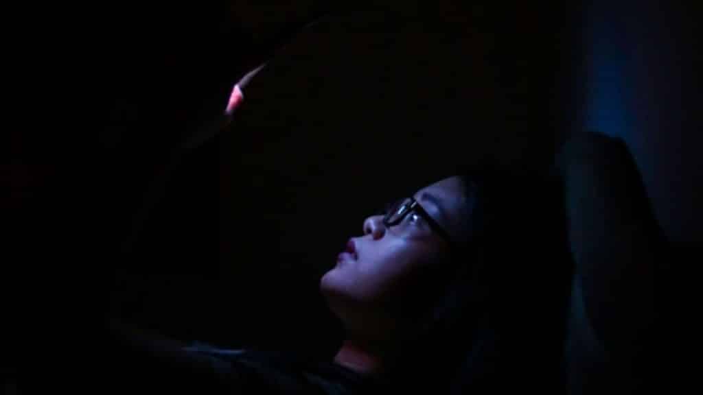 In a dark room, a person wearing glasses looks up at a glowing smartphone screen