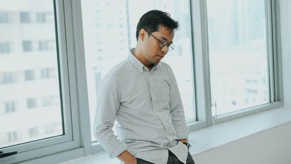 A man wearing glasses and a striped shirt stands by a window looking downward.