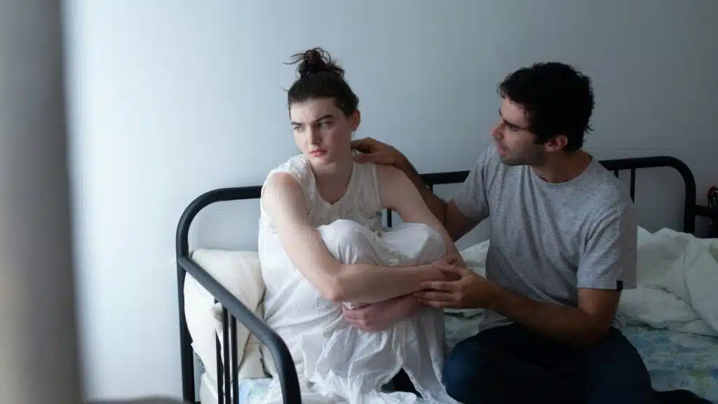 A woman sits curled up on a bed while a man places a hand on her.