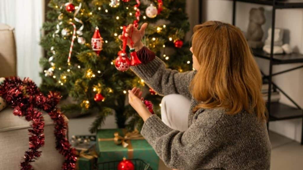 A woman in a grey sweater sits on the floor decorating a lit Christmas tree.