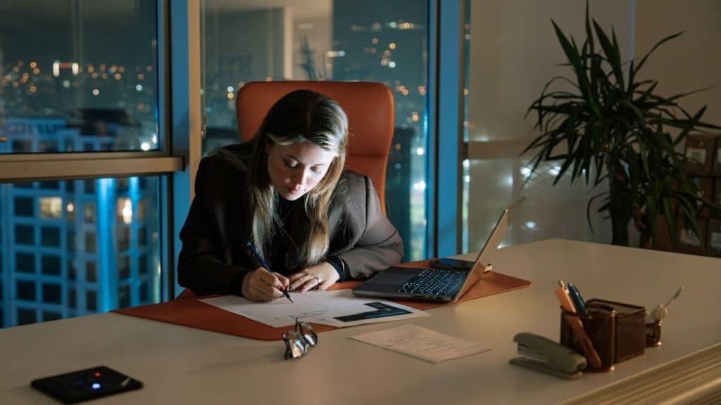 A woman sits at a desk in a high-rise office at night, writing on paper.