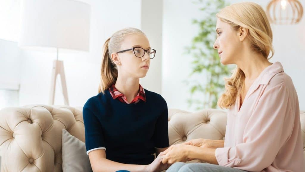 A woman sits on a tan sofa holding the hands of a young girl with glasses.