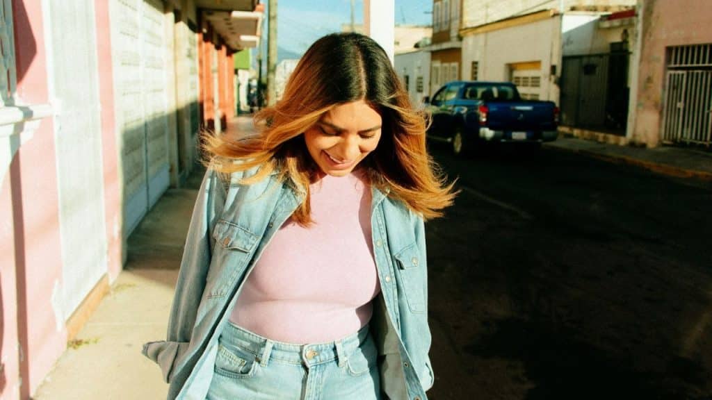 A smiling woman in a denim jacket walks down a sunny sidewalk past colorful buildings.
