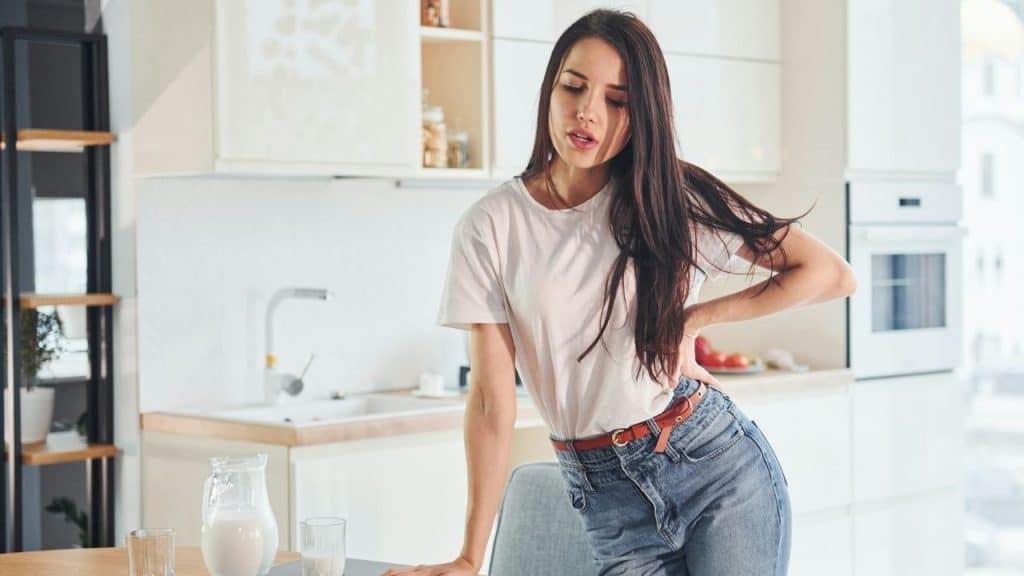 A woman in a white t-shirt and jeans stands in a kitchen holding her lower back.