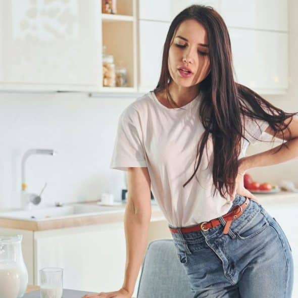 A woman in a white t-shirt and jeans stands in a kitchen holding her lower back.
