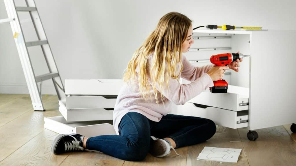 A woman sits on the floor using a red power drill to assemble white furniture.