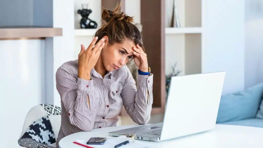 A woman sits at a desk with a laptop, holding her head with both hands.