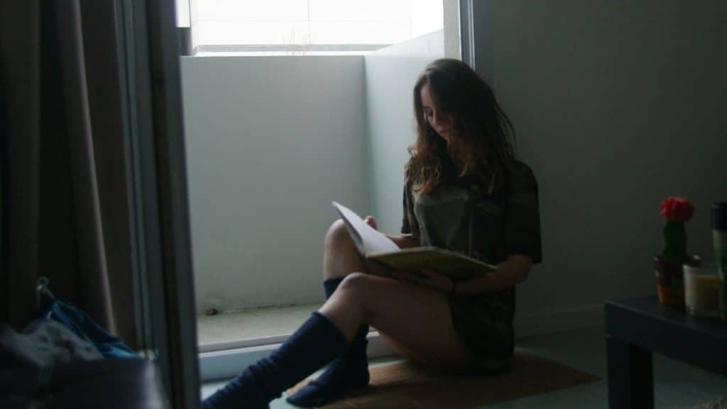 A woman in a camouflage shirt sits on the floor by a doorway reading a book