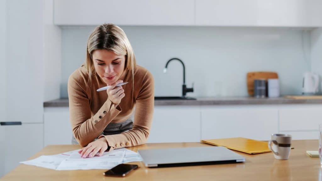 A woman leans over a wooden table in a kitchen reviewing several paper documents.