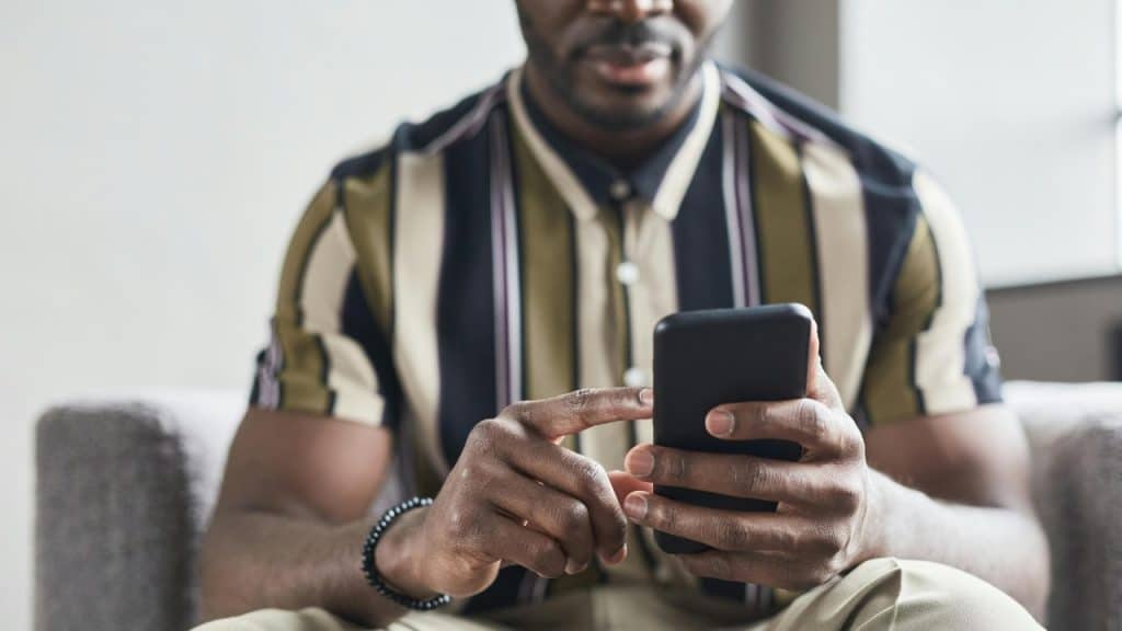 A man in a striped shirt sitting on a couch, holding and looking at a smartphone.