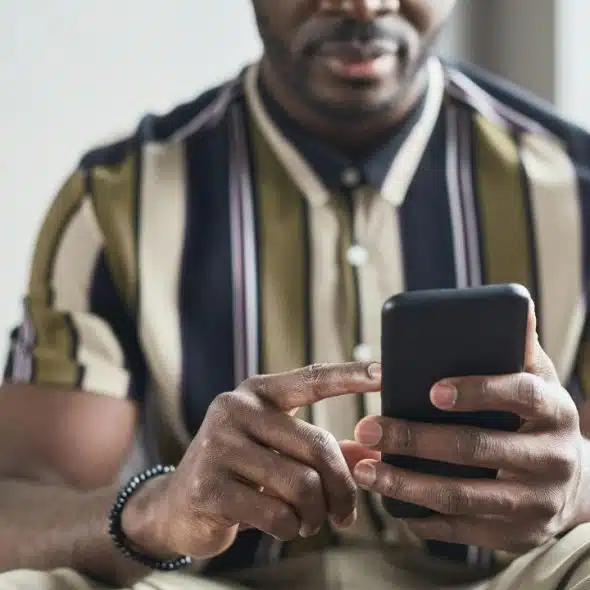 A man in a striped shirt sitting on a couch, holding and looking at a smartphone.