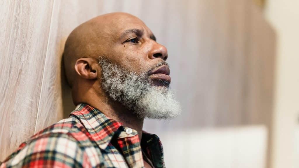 A bald man with a grey beard wearing a plaid shirt leaning against a wood wall.