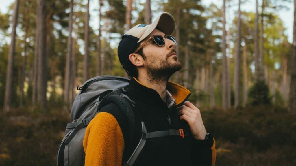 A man with a backpack, cap, and sunglasses looking up while standing in a forest.