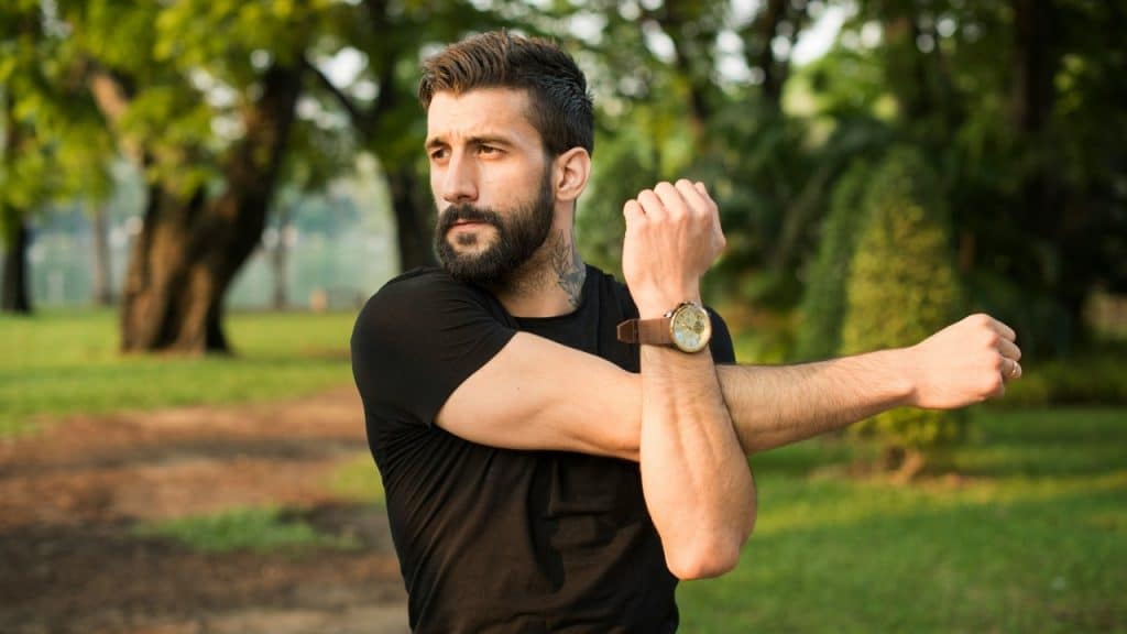 A man with a beard and neck tattoo stretching his arm in a green park setting.
