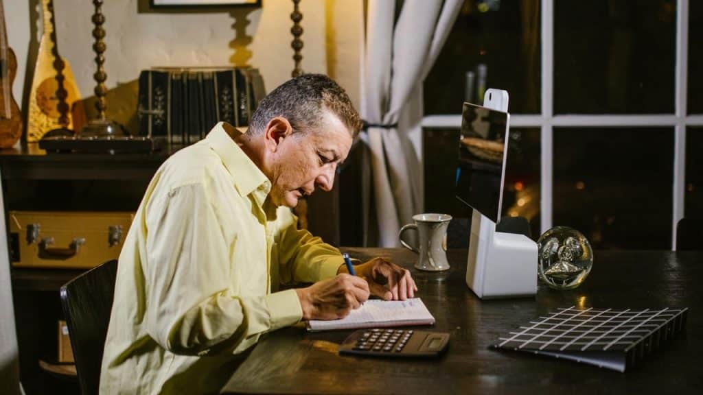 A man in a yellow shirt sitting at a dark desk writing in a notebook indoors.