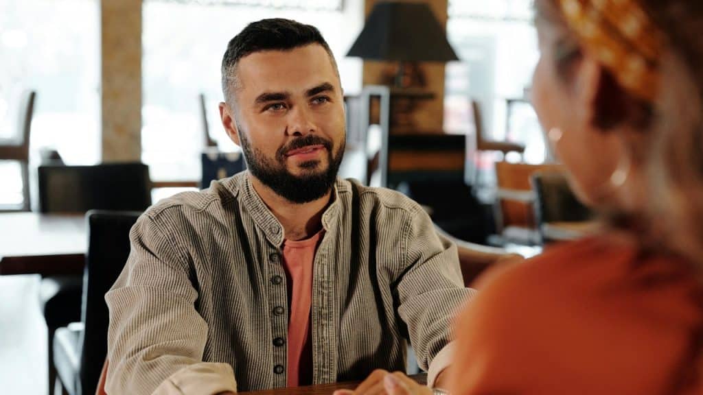 A bearded man in a beige shirt sitting at a table looking at a woman's face.