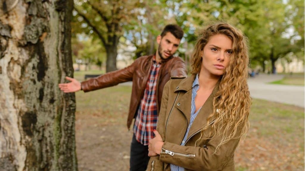 A woman with long wavy hair, wearing a brown jacket, is looking directly at the camera while a man gestures in the background near a tree.