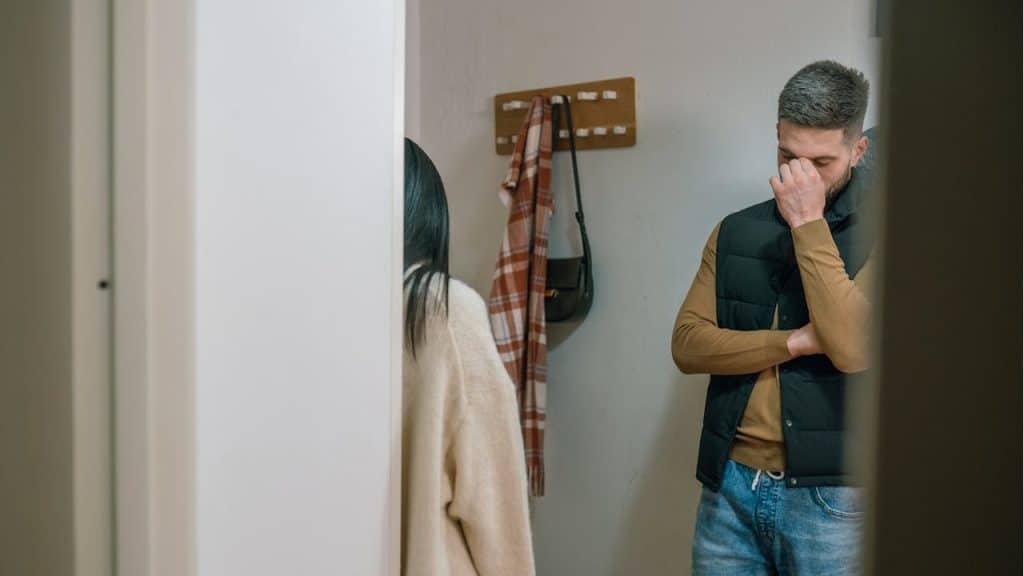 Man in a black vest and jeans holding his hand to his forehead while facing away from a person in a cream-colored sweater in a doorway.
