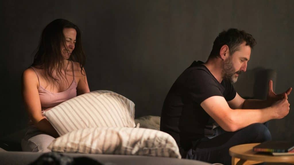 A woman smiling and holding a pillow next to a man who is looking down with a serious expression.