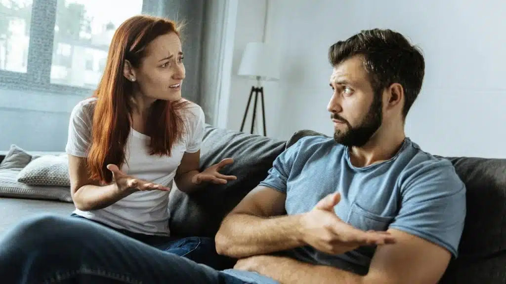 A woman and a man sit on a couch facing each other, both gesturing with their hands and appearing to argue or have an intense discussion in a living room.