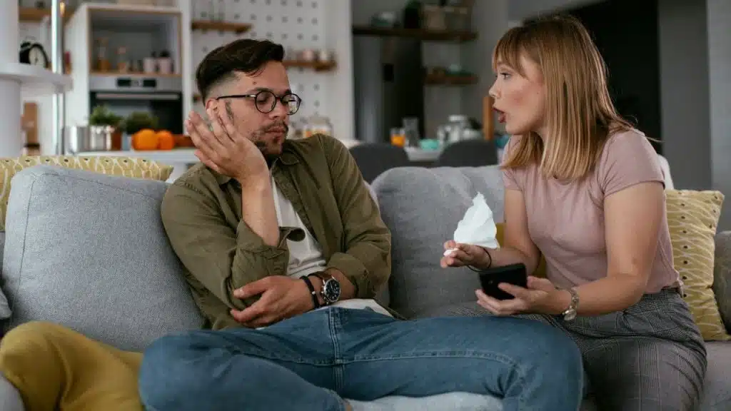A woman holds a phone and tissue while speaking to a man sitting beside her.