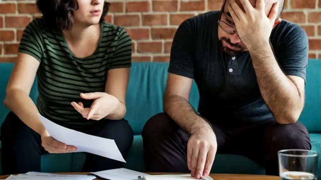 A woman holds a paper and gestures toward a man with his hand on head.