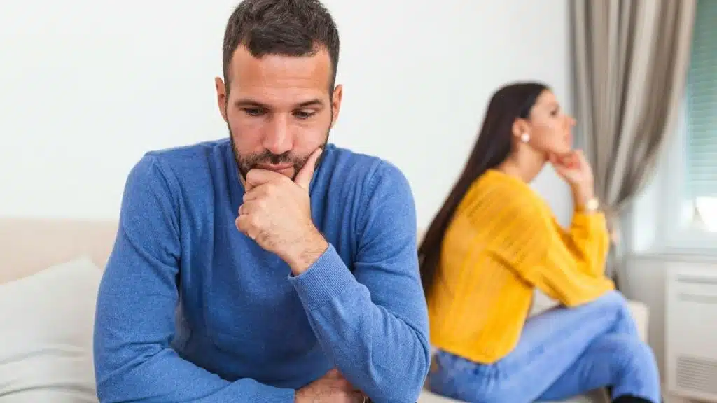 A man and woman sit on a couch looking in opposite directions with hands on chins.