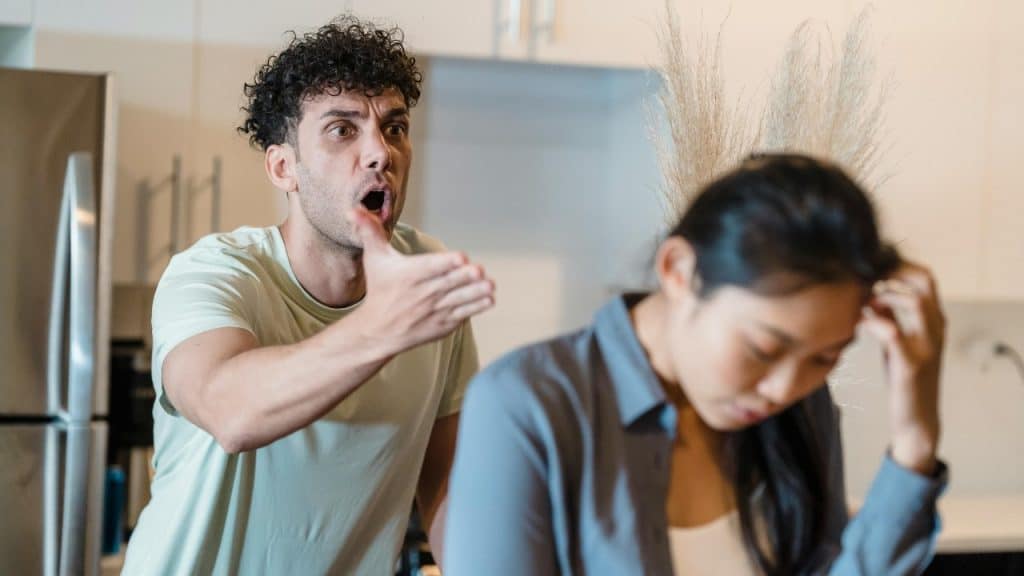 A man with curly hair gestures toward a woman who is looking down and away.