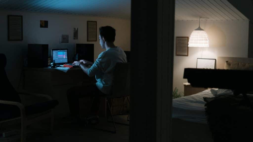 A person sits at a desk with a laptop and music equipment in a dark room.
