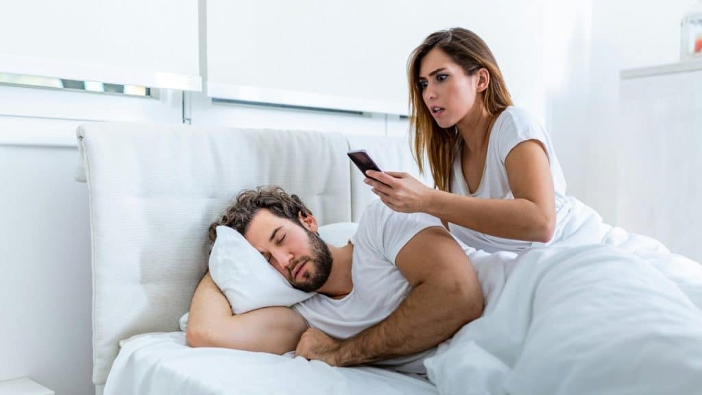 A woman sits on a bed holding a phone while looking at a sleeping man.