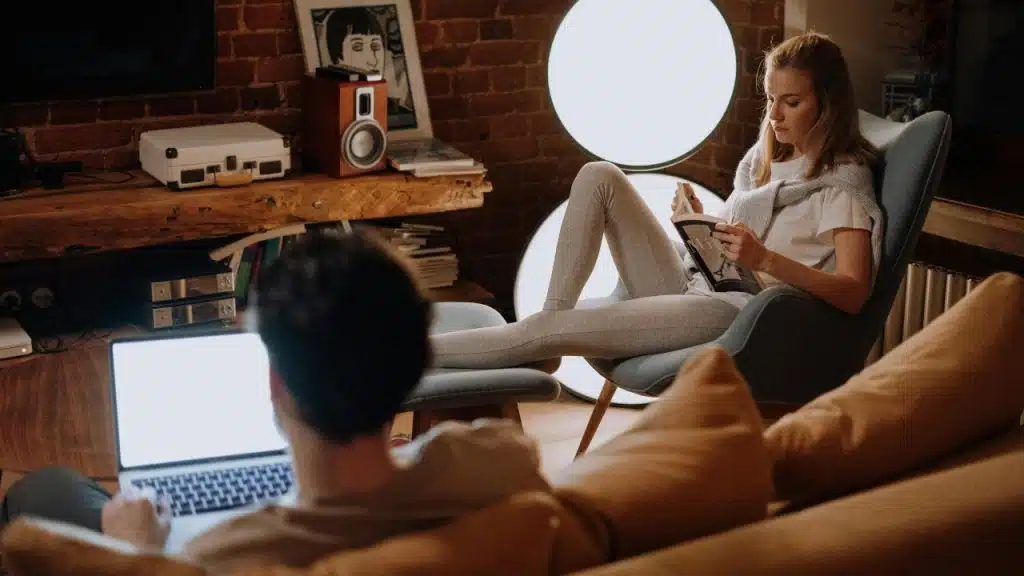 A woman reads a book in a chair while a man works on a laptop nearby.