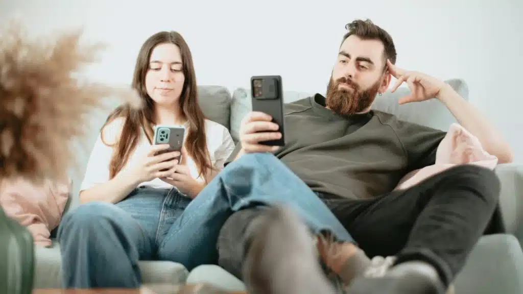 A man and woman sit on a couch together, both looking at their own smartphones.