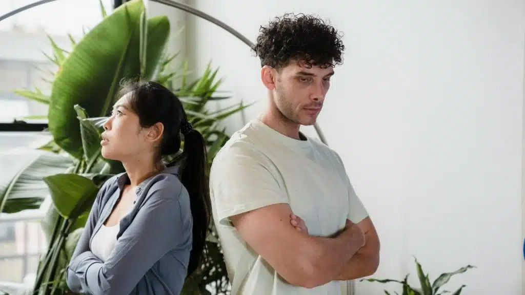 A man and woman stand back-to-back with their arms crossed in front of indoor plants.