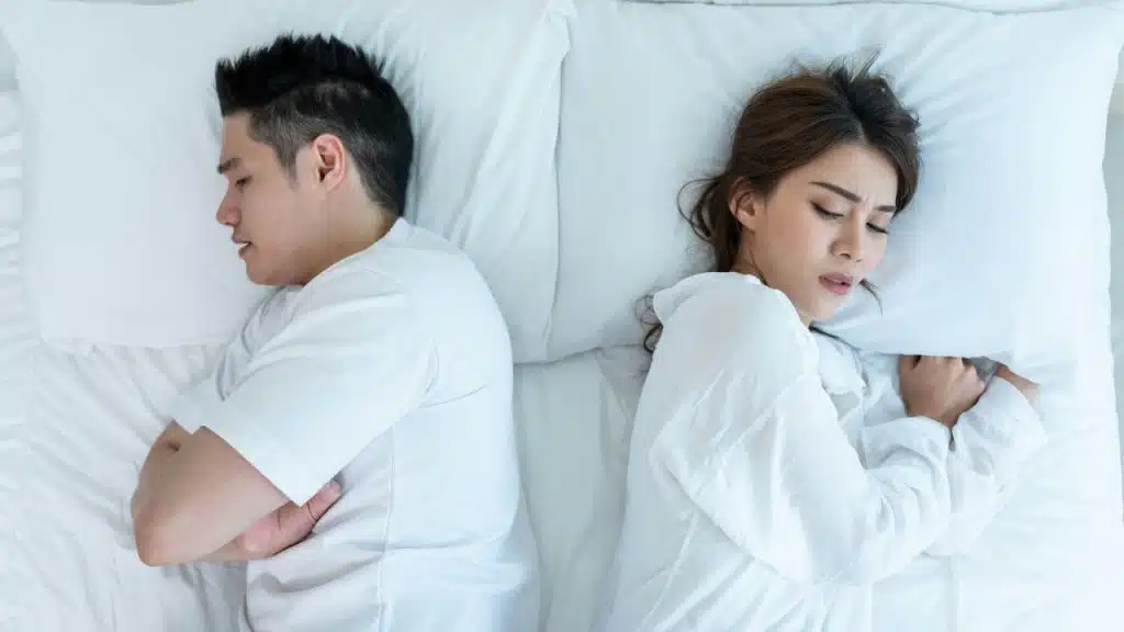 A man and woman lie in bed facing away from each other on white pillows.