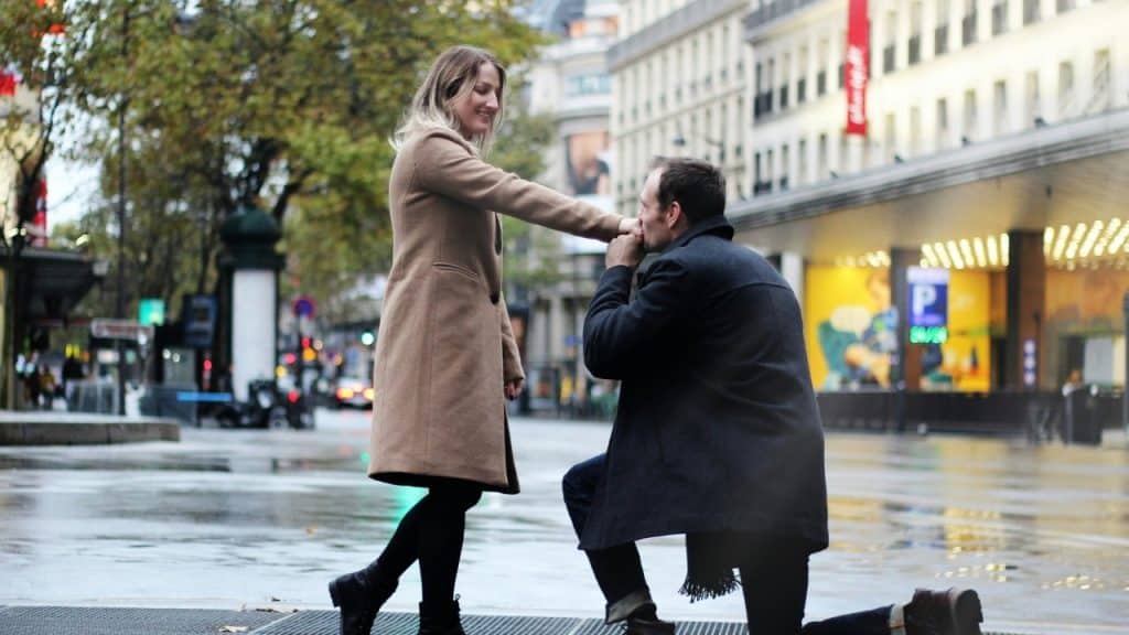 A man kneels on a wet city street and kisses a woman's hand.