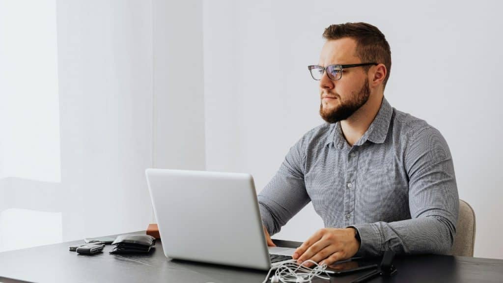 A man wearing glasses and a grey shirt looks at a laptop screen.