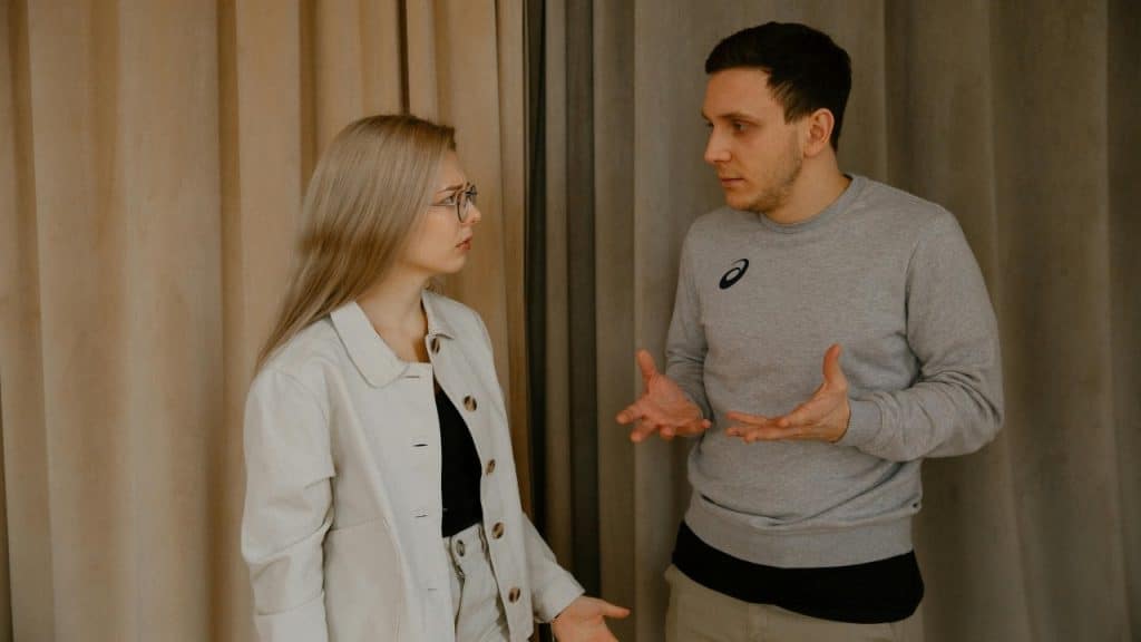 A man and woman stand near a curtain, both gesturing with their hands.