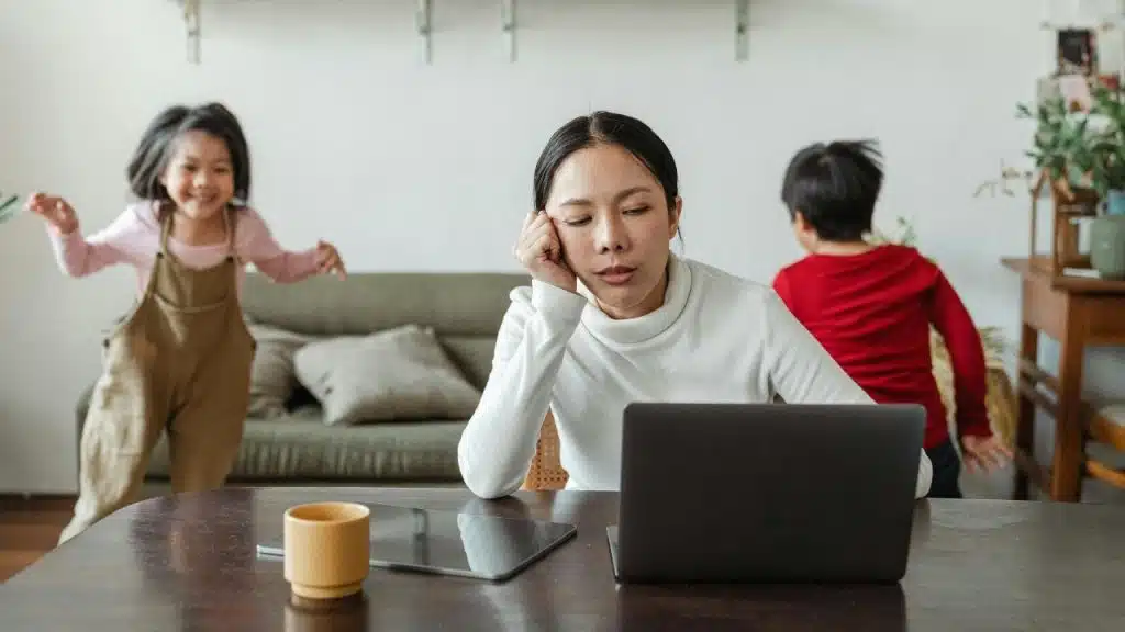 A woman sits at a table with a laptop while two children play behind.