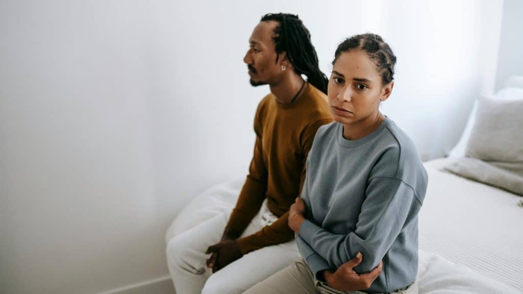 A man and woman sit on the edge of a bed looking in different directions.