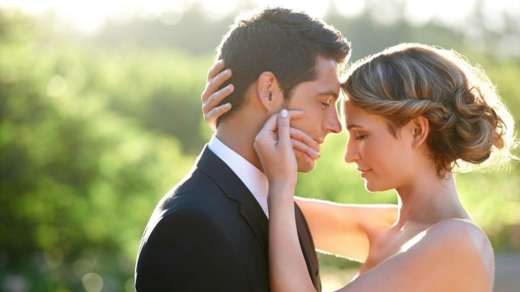 A man and woman in wedding attire stand close together with their foreheads touching.