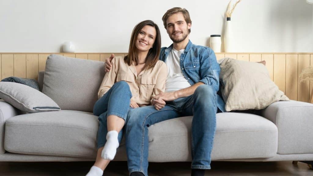 A man and woman sit together on a gray sofa in a neutral-toned living room.