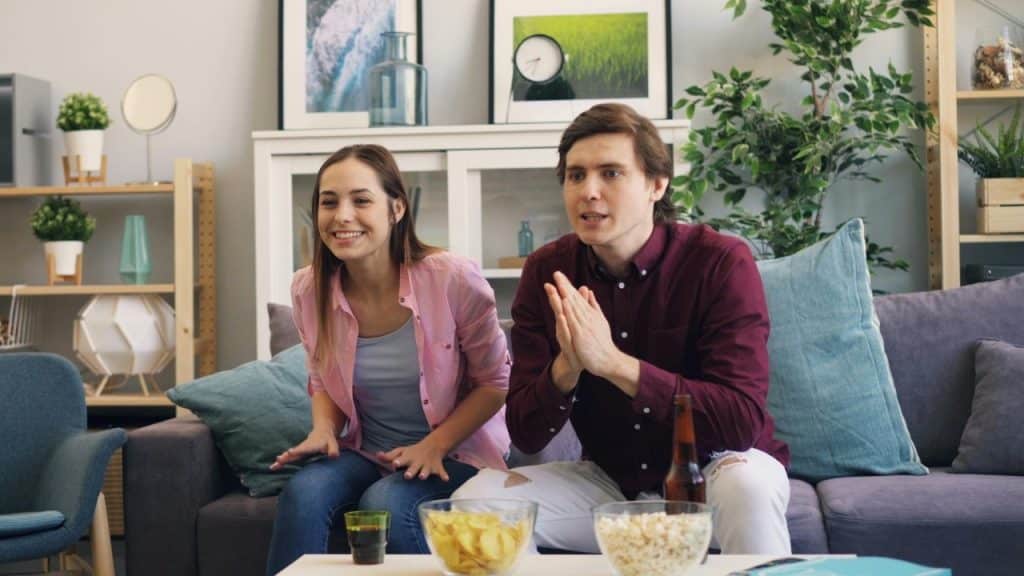 Two people sit on a purple couch watching a screen with snacks on the table.