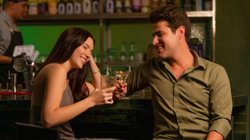 A man and woman smile at a dark bar while holding a cocktail and martini.