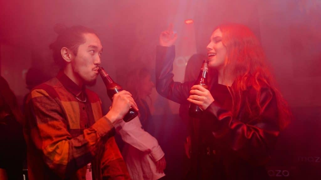 Two people hold glass bottles and dance in a dark room filled with red light.