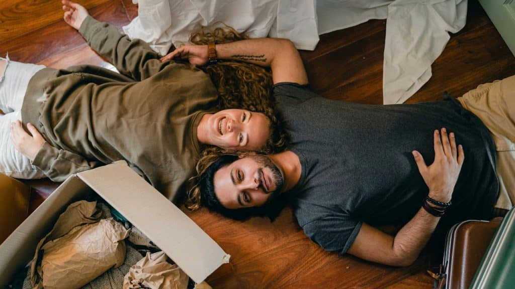 A man and woman lie on a wooden floor next to open cardboard moving boxes.