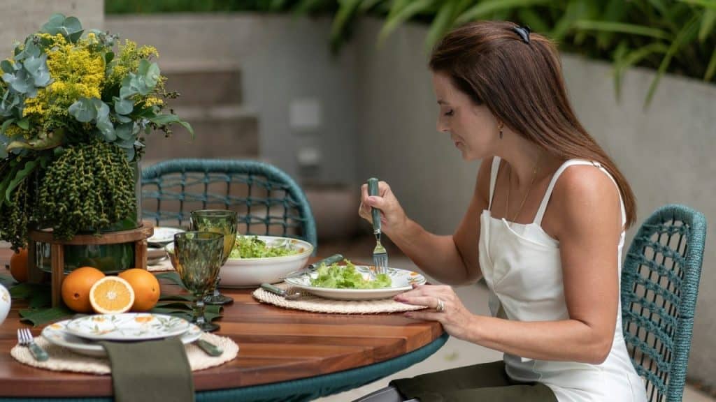 A woman in a white top eats a salad at a wooden outdoor dining table.